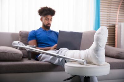 Man With Broken Leg Sitting On Sofa Using Laptop