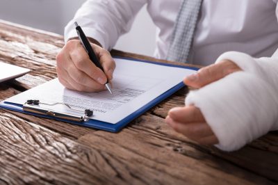 Close-up Of A Businessperson With Broken Arm Filling Health Insurance Claim Form On Wooden Desk